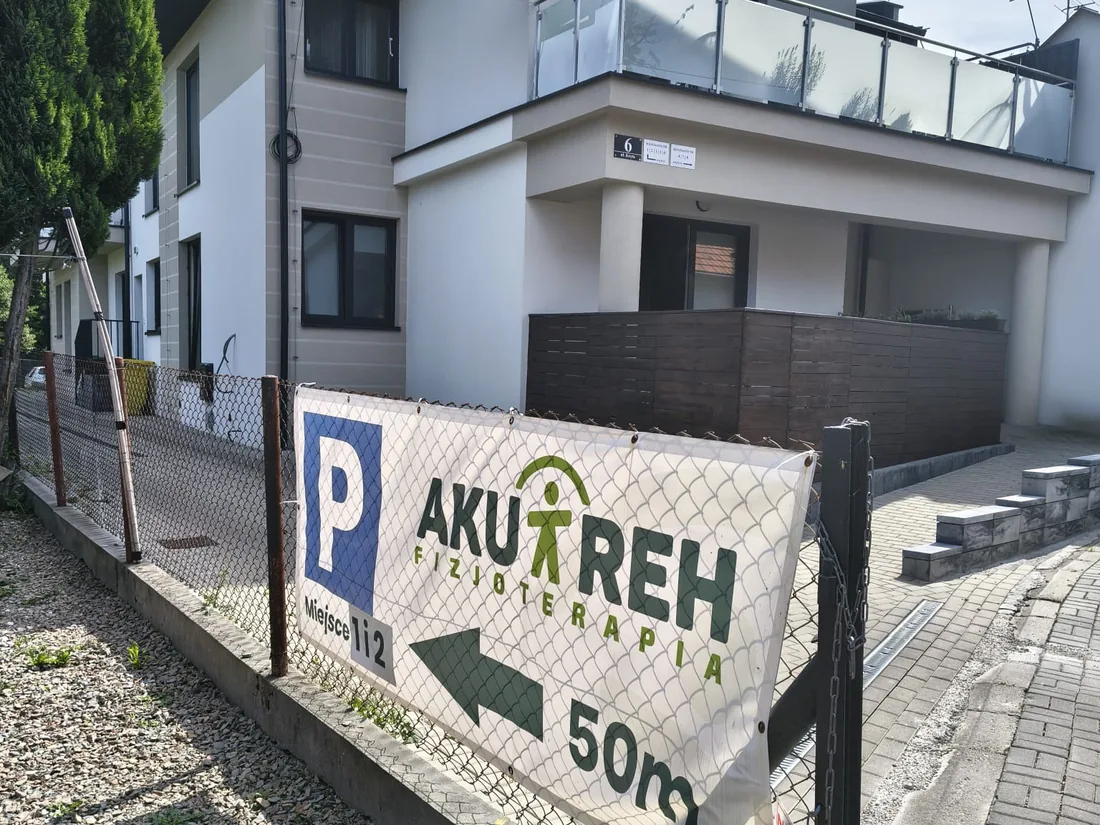 Baner P AKU-REH Fizjoterapia ze strzałką i napisem 50 m przy ogrodzeniu